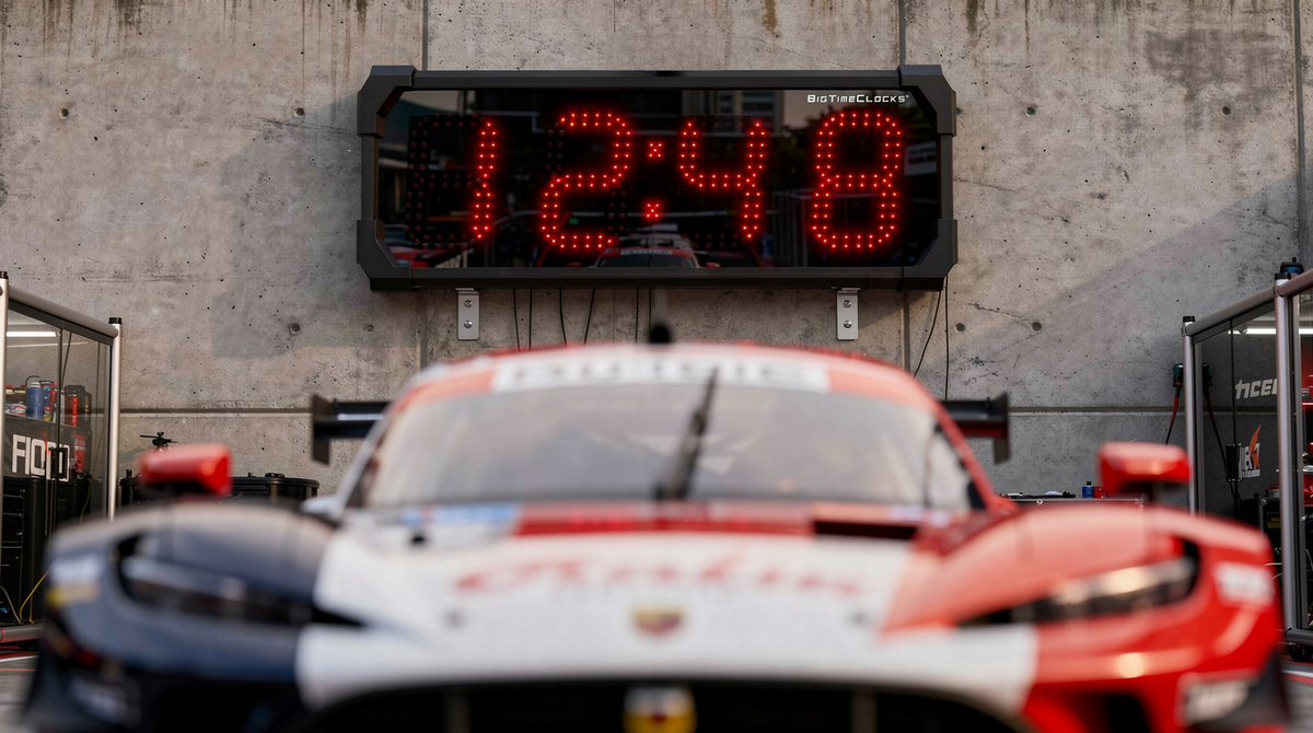 Race car in a pit lane with a BigTimeClocks digital clock displaying '12:48' on a concrete wall.